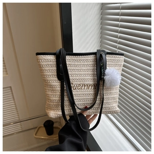 Detail of beige woven tote bag featuring black handles and a white fluffy pompom charm