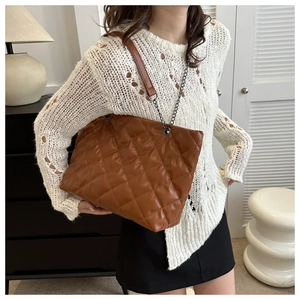 Brown quilted tote bag with metal chain strap worn by a model in a modern interior setting