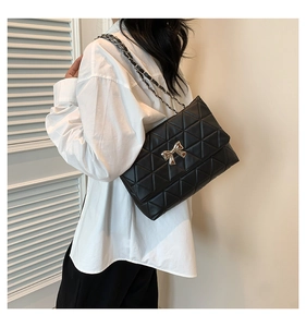 Detail of a black quilted shoulder bag with gold bow clasp worn by a model in a minimal setting