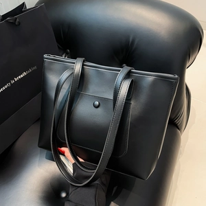 Minimalist large black tote bag in soft leather with a front snap pocket on a black sofa