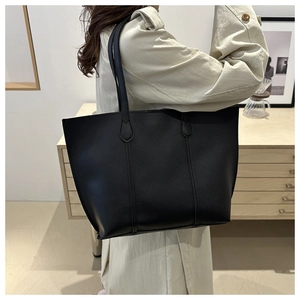 Minimalist large black tote bag on model shoulder with beige coat in home setting