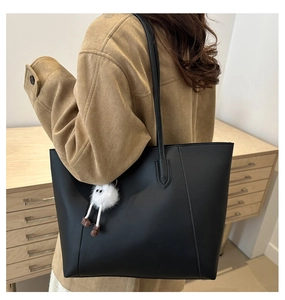 Model wearing large black faux leather tote bag with white plush charm and long straps