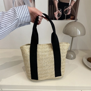 Trapezoid woven straw tote bag with black canvas straps on a minimalist desk