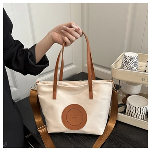 Close-up of cream canvas tote bag with brown leather handles and brand circular patch