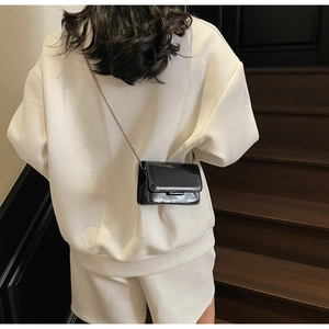 Detail of a black patent leather crossbody bag with a slim metal chain strap on a model