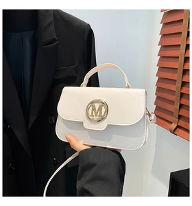 Detail of a creamy white crossbody bag featuring a golden circular M buckle on the flap