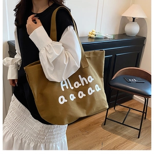 Detail of a large khaki canvas tote bag with white Alaha lettering used as a shoulder bag