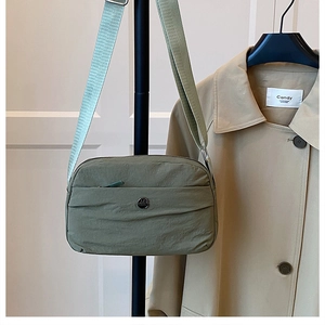 Detail of olive green nylon crossbody bag showing metal logo and wide strap on white wall