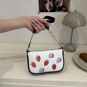 White shoulder bag with strawberry print and gold chain strap shown in a cozy setting