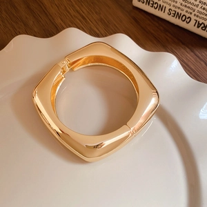 Polished gold square bangle with hinge closure on decorative plate and wooden table