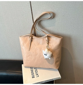 Detail of beige tote bag featuring embossed puppy patterns and a fluffy white pompom charm
