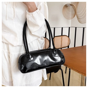 Glossy black baguette bag with long handles shown in a minimalist interior setting