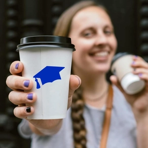 Close-up of a graduation cap sticker on a white coffee cup held by a smiling graduate