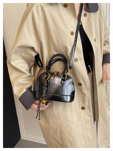 Detail of glossy black shell bag with gold hardware and bell charm worn on a trench coat