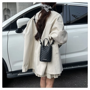 Detail view of black woven crossbody bag with metal plaque on beige coat