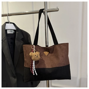 Detail of a brown suede tote bag with a golden triangular plaque and a plush bear charm