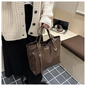 Detail of dark brown glossy tote bag with metal label held by a model in casual outfit