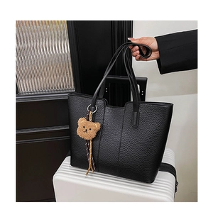 Detail of black pebbled leather tote bag with a brown teddy bear charm on a white suitcase