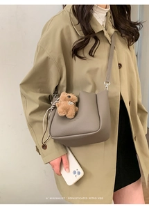 Grey leather bucket bag with teddy bear charm shown on a beige trench coat