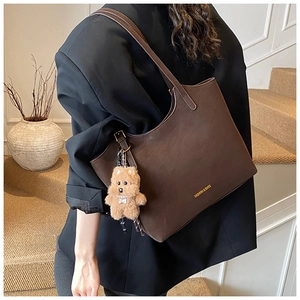 Brown leather tote bag worn on shoulder featuring a gold logo and teddy bear charm detail