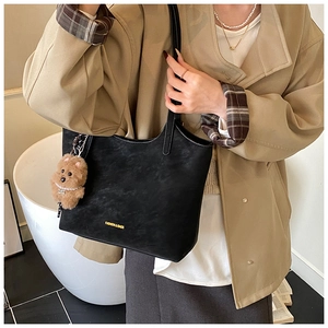 Model wearing a black matte leather tote bag with a brown plush bear charm and gold lettering