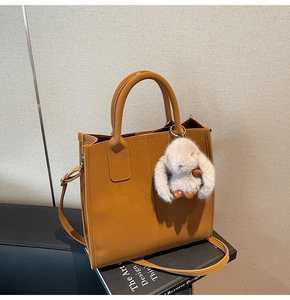 Detail of a brown leather tote bag with a plush rabbit charm on a minimalist background