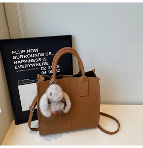 Brown tote bag detail with plush rabbit charm and adjustable shoulder strap on white desk