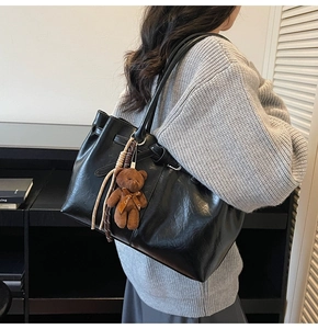 Detail of black glossy pleated tote bag with a brown plush bear charm worn by a model