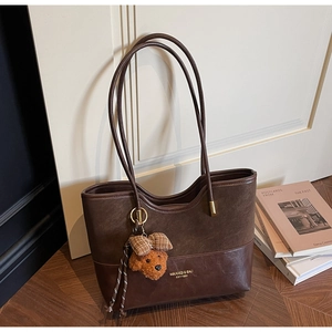 Detail of a brown leather tote bag featuring a plush bear charm on a warm wooden floor