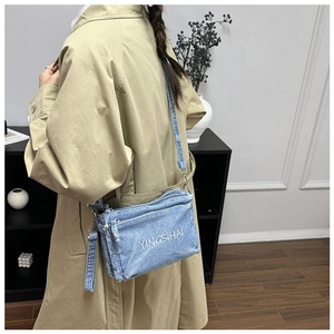 Detail of light blue denim crossbody bag with white embroidery and three zipper layers