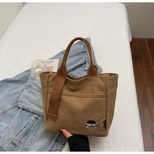 Detailed close-up of brown canvas tote bag with embroidered cartoon patch on white bedding