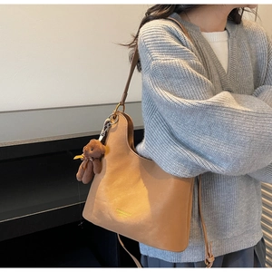 Detail der khakifarbenen Ledertasche mit Goldlogo und Bärenanhänger auf grauem Pullover