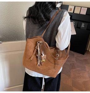 Brown leather tote bag worn by a model, featuring a cute bear plush charm detail