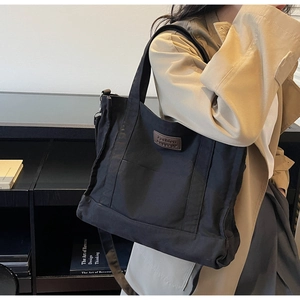 Model wearing a black canvas tote bag with adjustable shoulder strap and leather patch detail