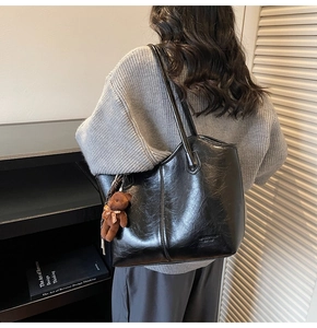 Detail of a large black glossy tote bag with a brown teddy bear charm on a model's shoulder