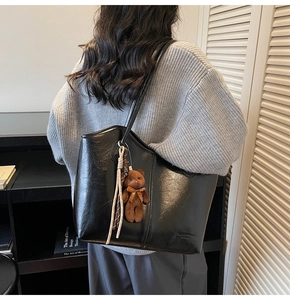 Detail of a large black PU leather tote bag with a brown teddy bear charm on model's shoulder