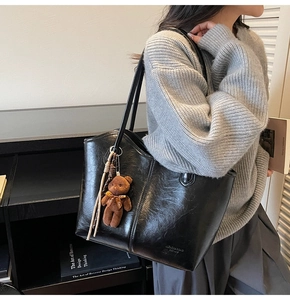 Detail of black glossy tote bag with slender straps and plush bear charm on a model