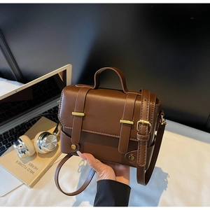 Detailed view of a brown box-shaped handbag with gold hardware on a desk with accessories