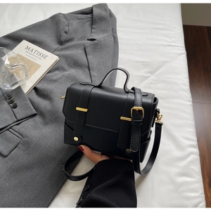 Detail of a black square crossbody bag with gold buckles on a white textured background