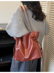 Brown leather bucket tote bag with chain strap and drawstring closure shown on shoulder