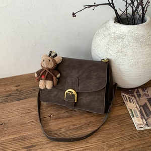 Detalle de bolso bandolera de cuero marrón oscuro con hebilla dorada y colgante de oso