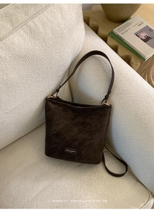 Detail der dunkelbraunen Wildleder-Achseltasche mit goldenem Logo auf einem Sofa