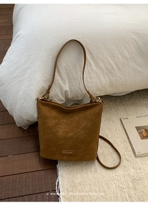 Detail einer braunen Wildleder-Beuteltasche mit Logo-Plakette auf Teppich und Magazin