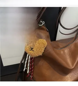 Close-up of a plush bear heart charm with woven cords on a brown leather shoulder bag