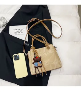 Detail of light khaki tote bag with a bear charm and long strap on a white cushion