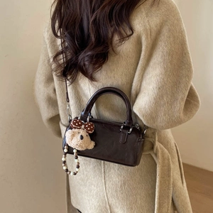 Deep brown cylinder crossbody bag with plush bear charm shown on a beige wool coat