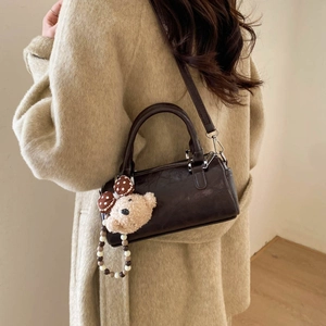 Deep brown Boston barrel bag with plush bear charm worn over a beige coat