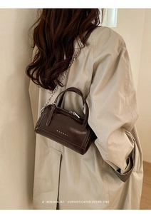 Detail of a dark brown Boston bag with chain strap worn over a beige trench coat