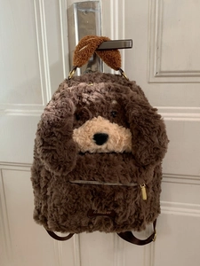 Cute brown plush puppy backpack with long ears and bone logo shown hanging on a door