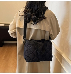 Detail of a black quilted crossbody bag with a wide strap shown on a beige trench coat